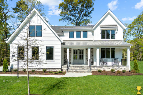 A 2-story Modern Farmhouse style residential building with white exterior walls, a black front door, and a gable roof, set on a grassy lot with trees in the background.