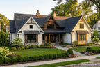 A large traditional-style house with white siding, dark gray shingled roofs, a covered front porch, stone accents, and a landscaped lawn with flowers and shrubs.