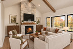 Great room with a stone fireplace, cream sofas, exposed ceiling beams and large windows overlooking autumn trees.