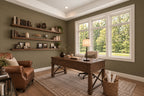 Willow Cove home office with olive-green walls, a wood desk, floating shelves and a brown leather armchair beside large windows.