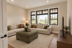 Home office/study furnished as a sitting room with a sectional sofa, green ottoman and wide black-framed windows.