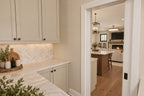 Walk-in pantry with cream cabinets, a pale countertop and herringbone backsplash, looking into the kitchen.