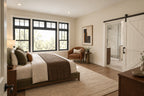 Main-floor primary suite with large windows, neutral bedding and a sliding barn door opening to the bathroom.
