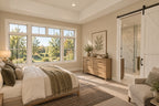 Main-floor primary suite with wide windows, neutral bedding and a sliding barn door opening to the bathroom.