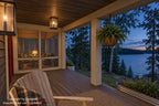 Dusk rendering of the covered front porch with a glowing wall lantern, rocking chair and lake beyond the porch.
