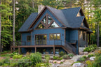Cottage rear exterior rendering with blue siding, a tall glazed gable, raised deck and walkout lower level in a wooded setting.