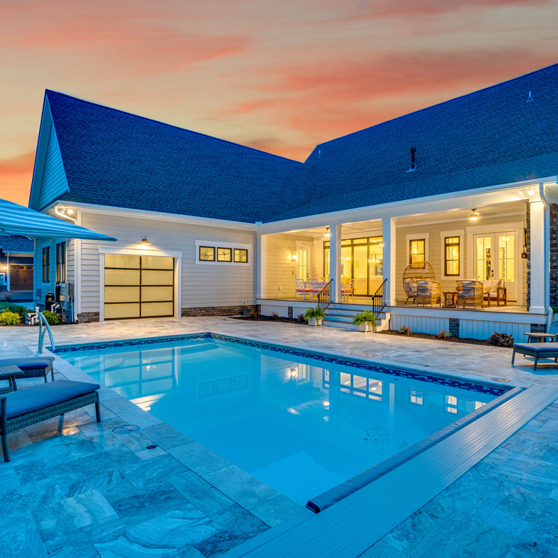 Custom home rear exterior by Royal Oaks Design Inc with in-ground pool, travertine deck, covered porch, and glass rear garage door at sunset